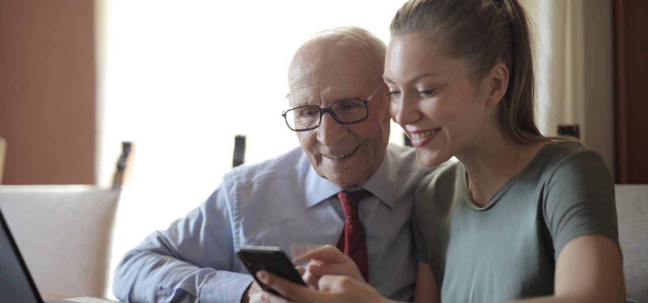 Smiling young woman in casual clothes showing smartphone to interested senior grandfather in formal shirt and eyeglasses while sitting at table near laptop