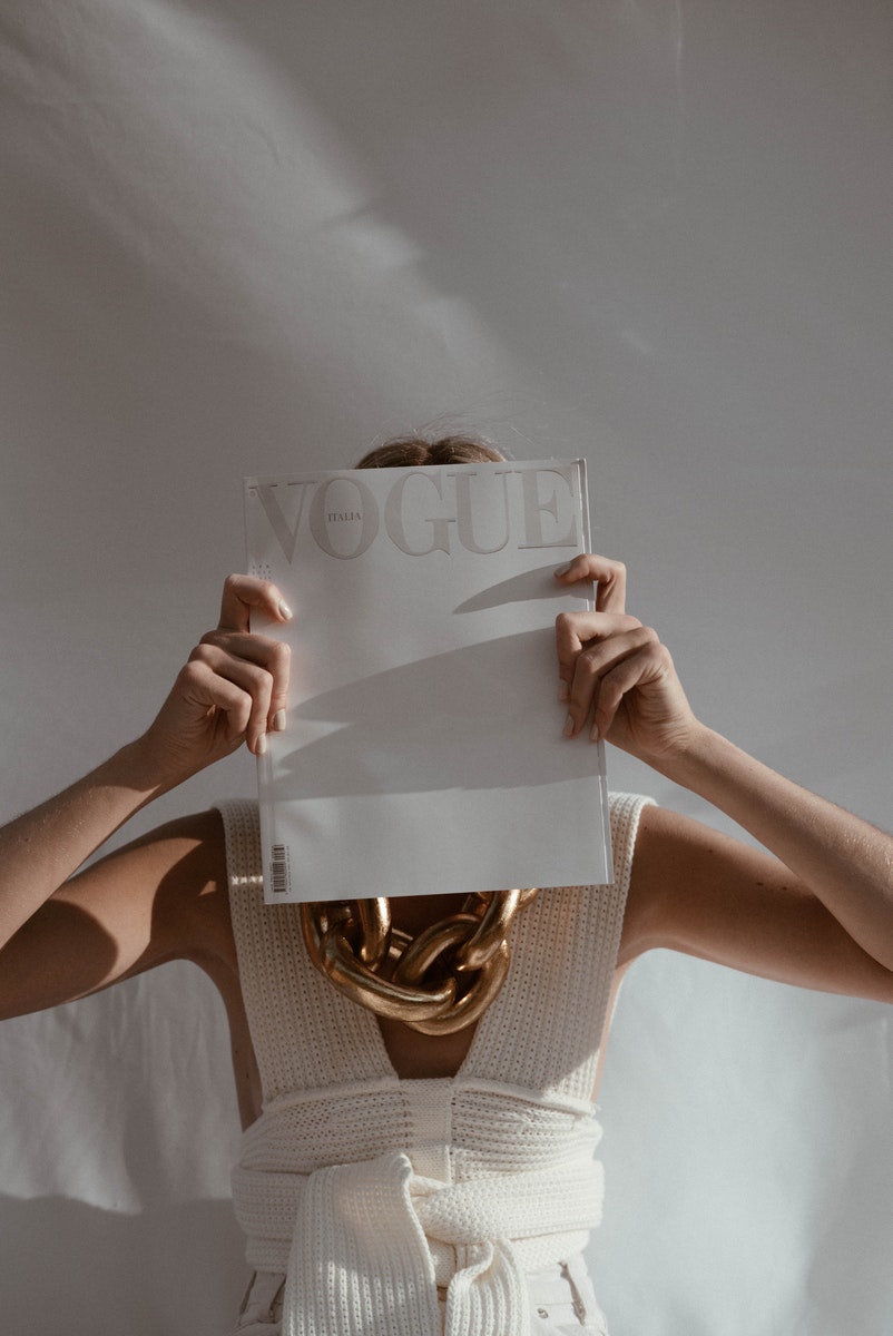 Unrecognizable female with big chain on neck standing on white background while covering face with fashion magazine in sunlight