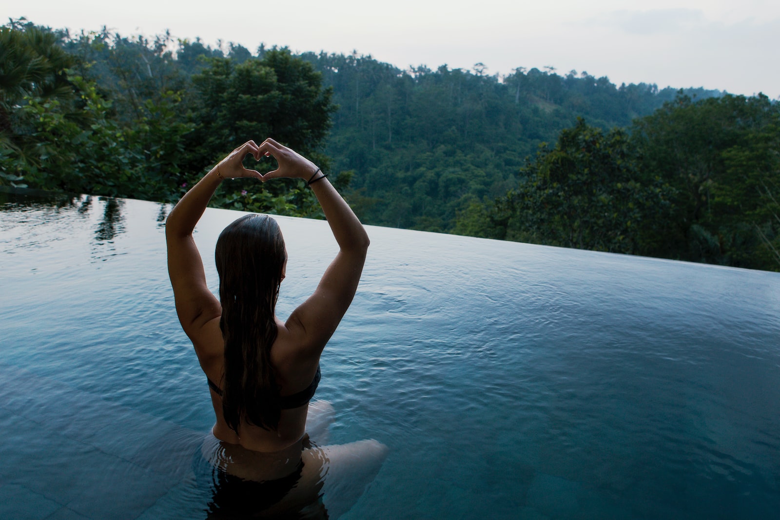 woman in infinity pool making heart hand gesture facing green leafed trees